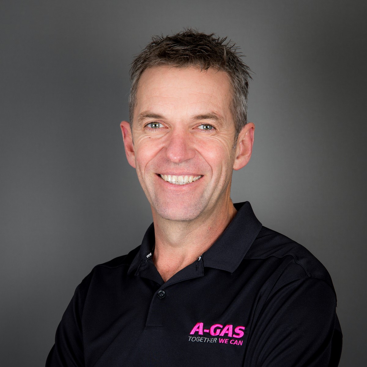 A man in a black polo with the pink A-Gas logo smiles at the camera with crossed arms