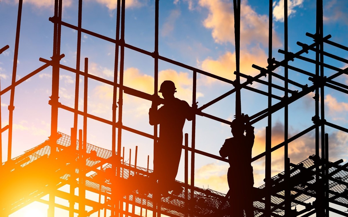A photograph of construction workers standing on scaffolding, silhouetted against a vibrant sunset.