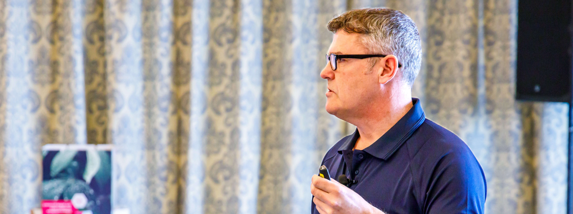A side-shot of a man in a blue polo and glasses delivering a presentation